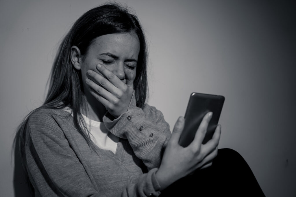 Sad desperate young teenager female girl on smart phone suffering from online bulling and harassment felling lonely and hopeless sitting on bed at night. CYberbullying and dangers of internet concept.