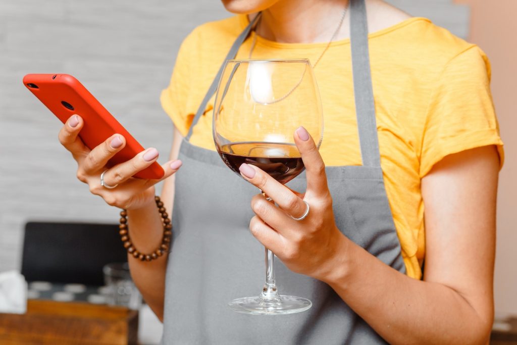 woman in apron looking at mobile phone and enjoying glass of red wine